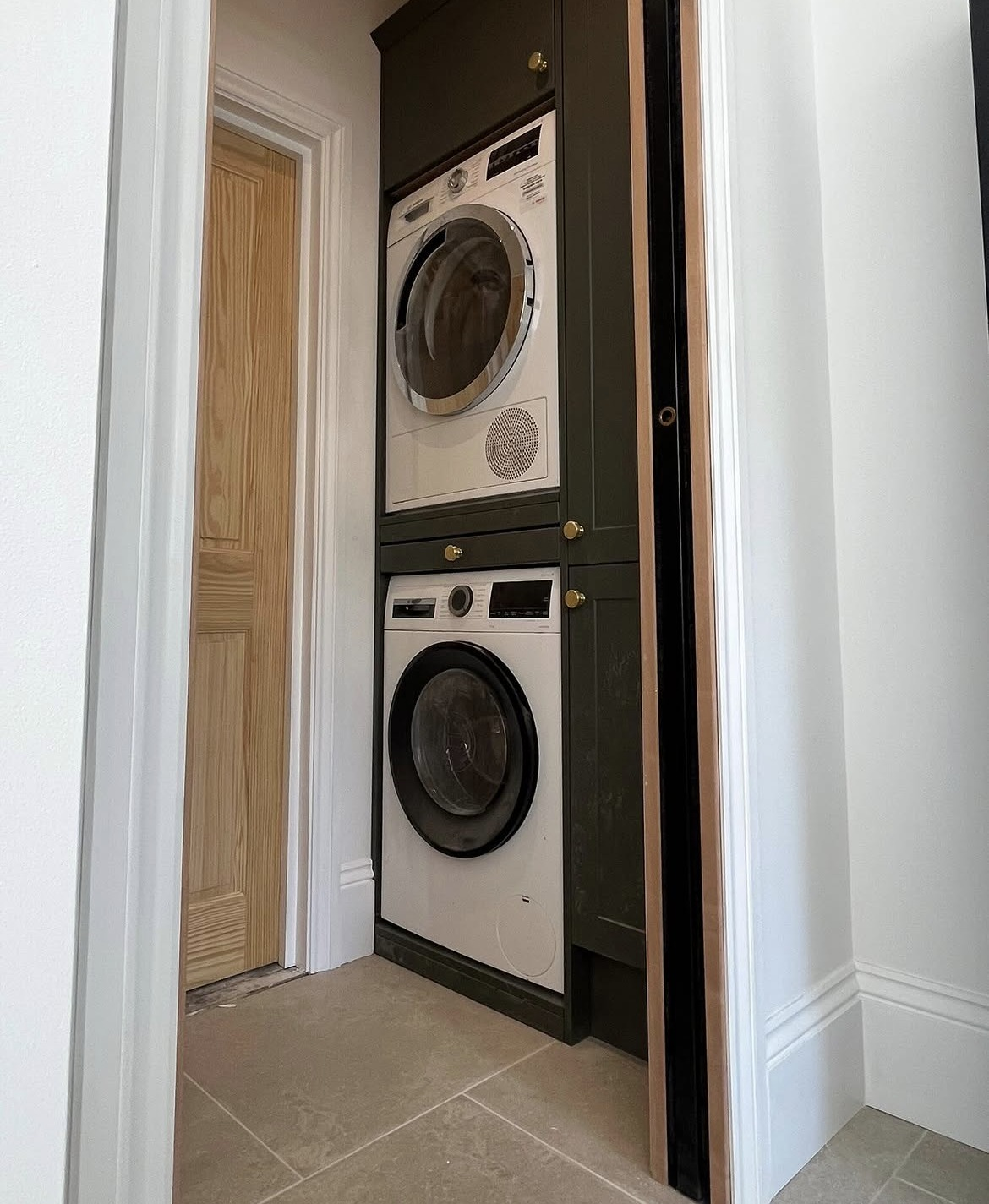 Bespoke joinery housing a stacked washing machine and dryer in a dark green fitted utility cupboard with brass handles